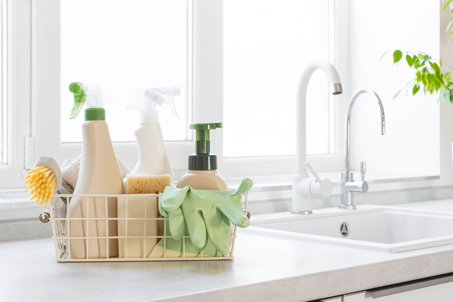 Cleaning products, brushes, sponges, wipes and rubber gloves in basket on kitchen counter near sink and window